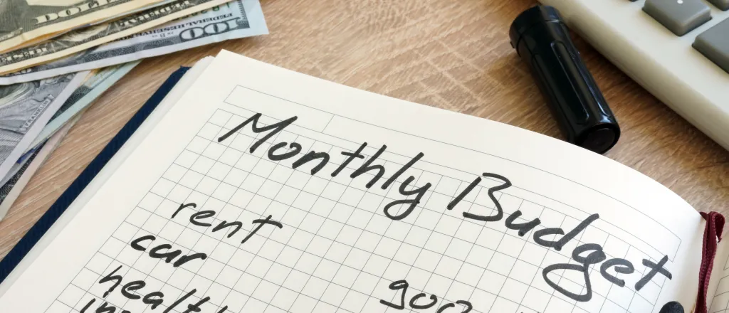 Notebook open to a page with hand-written "Monthly Budget" at the top. Cash and a calculator are nearby.