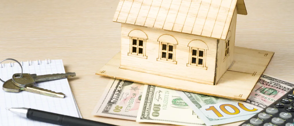 Model house made of wood resting on United State currency. A calculator, pen, notepad, and keys are nearby.