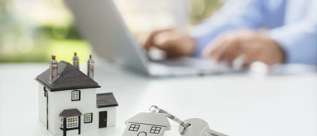 Miniature house and key on a keychain on a table. A person's hands and notebook computer are in the background.