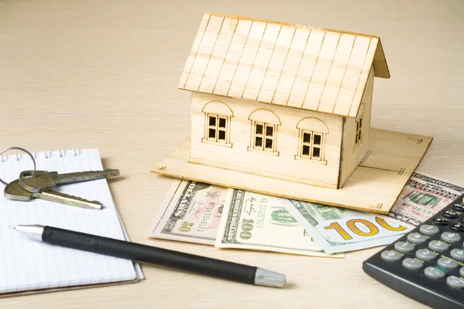 Model house made of wood resting on United State currency. A calculator, pen, notepad, and keys are nearby.
