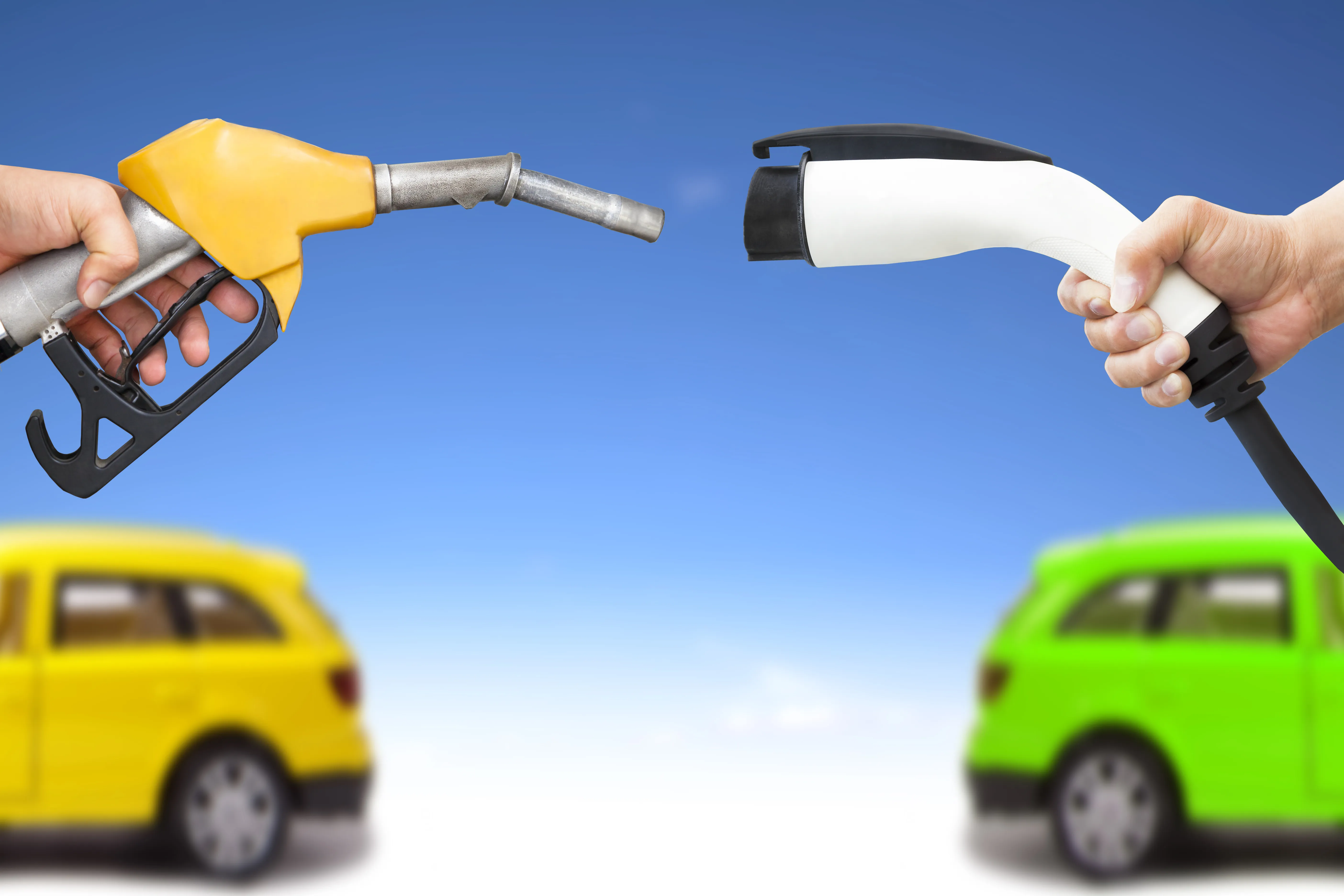 Hands holding a gasoline fuel nozzle and electric charging cable in the foregrounds, with two vehicles in the background.
