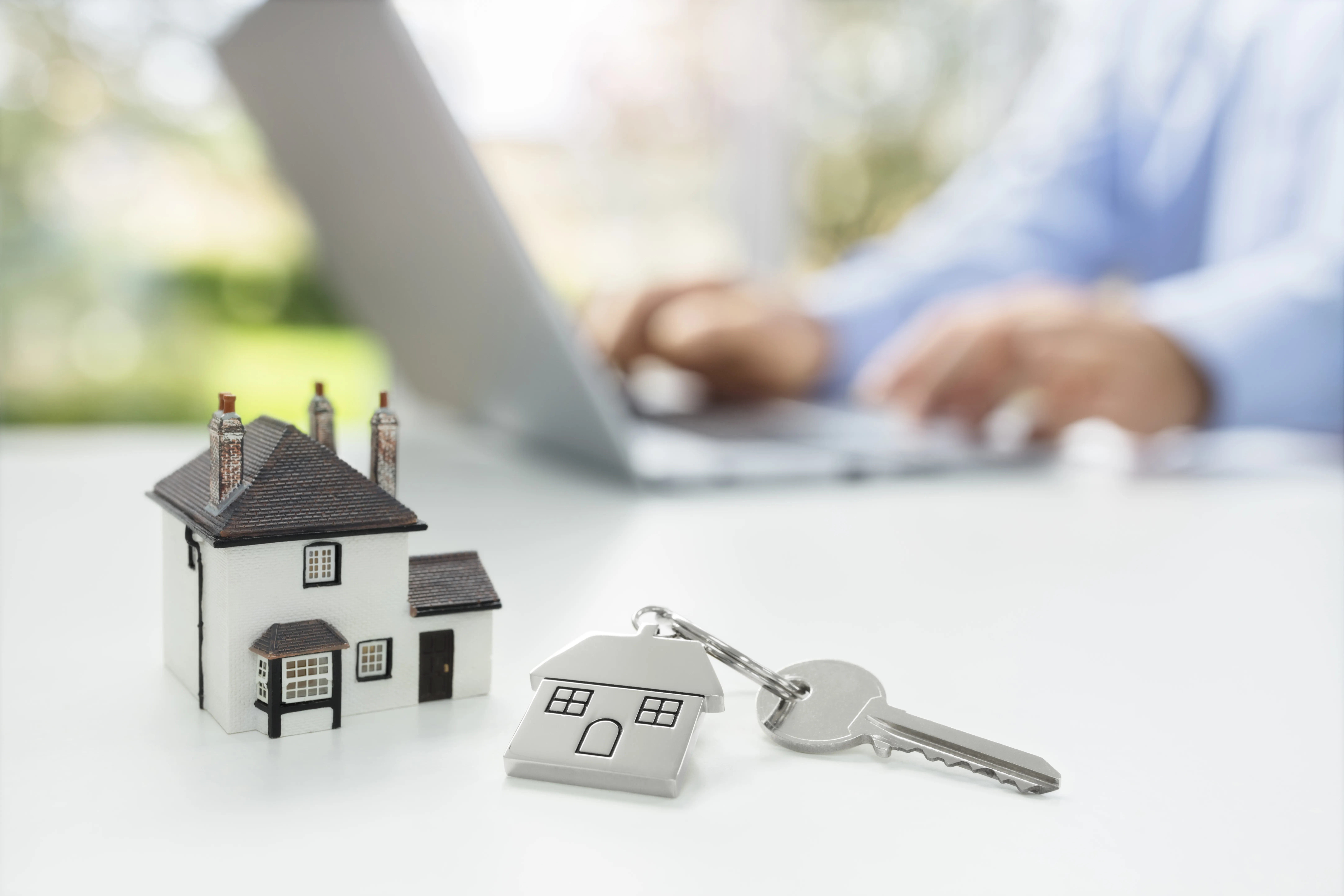 Miniature house and key on a keychain on a table. A person's hands and notebook computer are in the background.